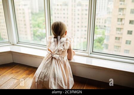 Elegante bambina con capelli biondi seduta a casa vicino alla finestra durante l'auto isolamento del coronavirus covid-19 Foto Stock