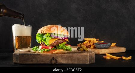 Panorama di Burger e birra. Hamburger con manzo, formaggio, cipolla, pomodoro e insalata verde, vista laterale su sfondo scuro con una p Foto Stock