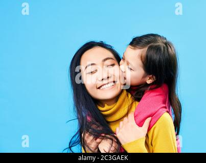 Piccola figlia carina abbracciare e baciare la sua madre felice isolato su sfondo blu Foto Stock
