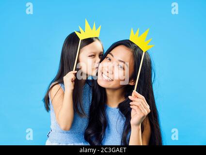 Carino piccola figlia baciare la sua mamma felice giovane tenendo carta corona su bastone su sfondo blu divertirsi insieme Foto Stock