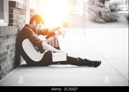 Solo maschio seduto da un muro di mattoni con una chitarra E la Bibbia su questo giro Foto Stock