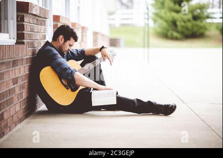 Solo maschio seduto da un muro di mattoni con una chitarra E la Bibbia su questo giro Foto Stock