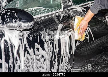 Primo piano della parte anteriore di una moderna auto nera. Sfregamento e pulizia con una mano maschile su un autolavaggio. Foto Stock
