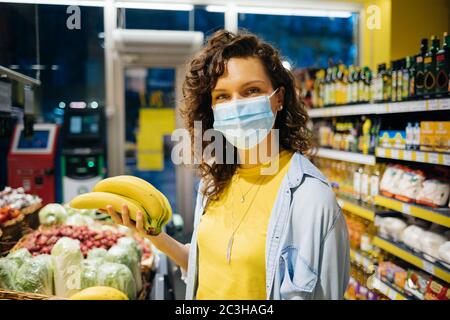 Giovane donna che indossa una maschera medica protettiva in piedi in negozio di alimentari che tiene mazzo banane guardando la macchina fotografica. Foto Stock