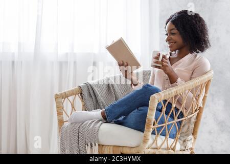 Passatempo del fine settimana. Giovane donna Nera rilassante in poltrona con libro e caffè Foto Stock