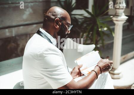 Vista laterale di un uomo africano maturo e calvo con barba nera e pulita, in occhiali ed elegante vestito bianco seduto su una veranda, illuminato dal sole Foto Stock