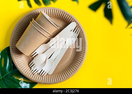 utensili da cucina in legno e carta utensili ecologici riutilizzabili vuoti - piatti, tazze e forcelle su sfondo giallo con foglia verde tropicale, Foto Stock