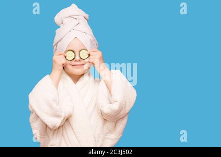 Divertente bambina in camice bianco e asciugamano con maschera per la pelle viso e cetrioli su occhi isolati su sfondo blu. Trattamenti per la cura della pelle Foto Stock