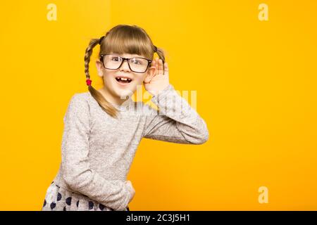 Giornalista bambina in occhiali con sorpresa euforia viso e simpatici pigtail ascoltando qualcosa che tiene la mano all'orecchio isolato su yel Foto Stock