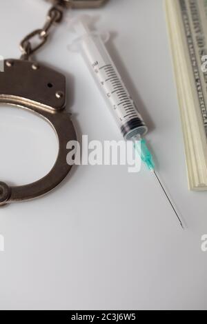 Immagine verticale ad alto angolo di manette, una siringa e alcune bollette del dollaro su una superficie bianca Foto Stock