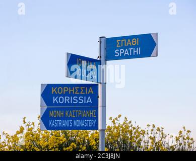 Grecia, Isola di Kea. Segnaletica stradale bilingue, greca e inglese su palo, cielo blu e sfondo giallo fiori selvatici. Indicazioni stradali su un incrocio. Foto Stock