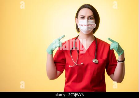 Ritratto di bella donna medico con stetoscopio che indossa scrub rossi, indossa una maschera protettiva, con le dita puntate posando su un isolato giallo Foto Stock