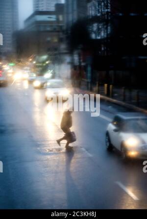 Immagine sfocata verticale di una persona che attraversa una strada con auto che guidano intorno in una giornata piovosa Foto Stock