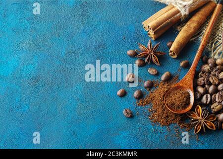 chicchi di caffè sparsi su fondo di tessuto blu, stelle di anice, bastoncini di cannella e caffè macinato in un cucchiaio di legno. Vista dall'alto Foto Stock