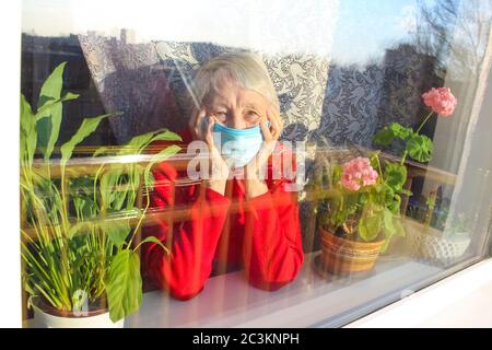 Il Covid-19, salute, sicurezza e pandemia concetto - anziana donna solitaria indossare maschera medica protettiva seduta vicino alla finestra a casa per la protezione dal virus Foto Stock