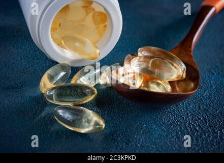 Capsule di gelatina gialla con un piccolo farmaco Omega-3, in un cucchiaio di legno, su fondo testurizzato blu scuro. Foto Stock