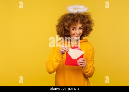 Messaggio d'amore. Ritratto di felice donna angelica riccia con santa nimbus che tiene lettera in busta e guarda la macchina fotografica con un sorriso giocoso. indo Foto Stock