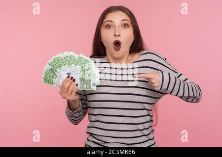 Ritratto di felicissima donna bruna in felicissima felpa a righe gridando in eccitazione e puntando banconote in euro, scioccato dalla vittoria della lotteria, grande j Foto Stock