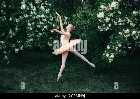 Ballerina in un tutu bianco che danzano e mostrano le pose classiche del balletto e saltano in alto all'aperto. Sfondo fiori verdi Foto Stock