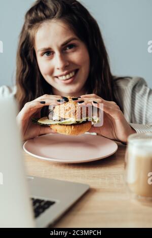 Ritratto di giovane ragazza adulta che lavora da casa mangiare Burger, Freelance concetto di lavoro remoto Foto Stock