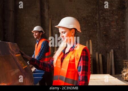 Operatrice professionale di falegnami in officina. Operatori che indossano elmetti, abiti e guanti protettivi. Foto Stock