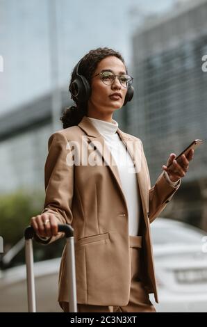 Donna d'affari in piedi sulla strada con bagagli e telefono cellulare. Donna in viaggio d'affari in attesa del suo taxi per arrivare. Foto Stock