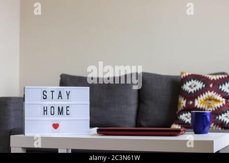 Scrivania con un computer portatile e una tazza di caffè, con un pannello che dice 'Stay home - Covid-19 concetto Foto Stock