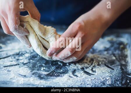 Un uomo in un grembiule blu sta impastando l'impasto con le mani su una teglia scura cosparsa di farina. Cottura. Torta fatta in casa. Foto Stock