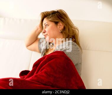 Donna bionda che si stendeva su un divano coperto da una coperta rossa con la mano sulla testa con un'espressione annoiata Foto Stock