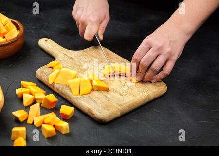 Persona che taglia una zucca su tagliere di legno su tavola di pietra. Mani femminili con coltello in acciaio, pezzi di zucca Foto Stock