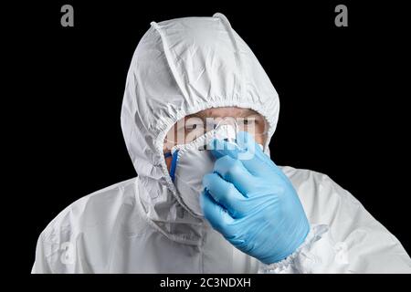 Uomo vestito con tuta protettiva bianca, respiratore e occhiali isolati su sfondo nero. La persona corregge la maschera del respiratore con la mano guanto. Personale Foto Stock