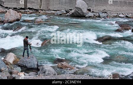 Fotografo di viaggio sul pericoloso fiume allagato in Azad Kashmir. In piedi su una roccia e catturare l'attimo con la sua fotocamera sul treppiede. Foto Stock