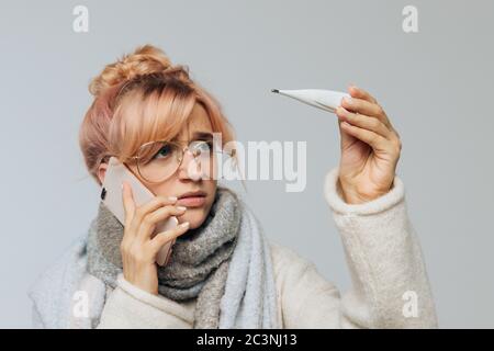 Ritratto closeup di donna europea malsana in bicchieri presi freddo e guardando termometro. Malata giovane bionda femmina in sciarpa sentirsi male, parlare Foto Stock