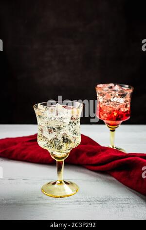 Un bicchiere con acqua frizzante ghiacciata in primo piano e un bicchiere con acqua frizzante ghiacciata e sciroppo rosso sullo sfondo. Tavolo bianco in legno, gr. Scuro Foto Stock