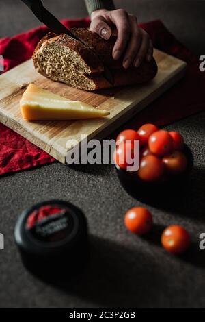 Donna che affetta pane di segale su un tagliere di legno. Un pezzo di parmigiano, pomodori ciliegini, un piccolo camioncello di formaggio in primo piano. Moody nat Foto Stock