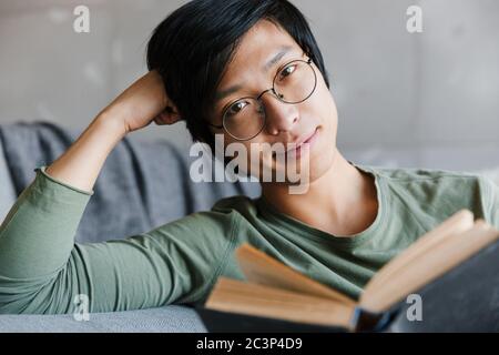 Immagine di bel giovane asiatico che indossa occhiali leggere libro mentre si siede sul divano in appartamento Foto Stock