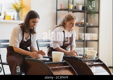 Due artisti femminili di ceramica in grembiule che usano materiale di argilla che lavora in studio di vasaio. Formazione a distanza freelance serale. Avventura hobby per adulti. Inverno Foto Stock
