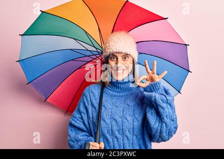 Giovane bella donna che tiene ombrello colorato facendo ok segno con le dita, sorridente amichevole gesturing eccellente simbolo Foto Stock