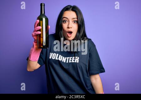 Giovane donna bruna indossa t-shirt volontario picking bottiglia di vetro su sfondo viola spaventato in shock con un viso a sorpresa, paura ed eccitato wi Foto Stock