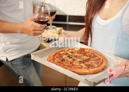 Immagine tagliata di giovane donna che porta vassoio con pizza appena appoggiata al tavolo quando il suo ragazzo porta bicchieri di vino rosso Foto Stock