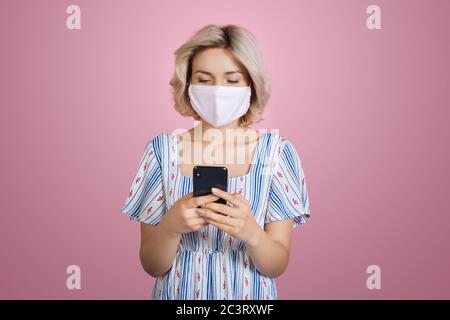 Bella donna bionda in maschera medica che indossa un bel vestito è chiacchierare su mobile in posa su una parete rosa studio Foto Stock