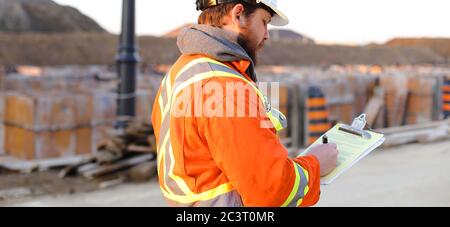 Banner di ispettore in abbigliamento da lavoro arancione con blueprint sul cantiere. Foto Stock