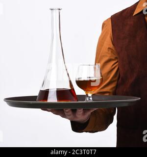 Cameriere con bicchiere e bottiglia di whisky sul vassoio. Concetto di bevande del ristorante. La mano dei maschi tiene il cognac su sfondo bianco. Il barman in pelle scamosciata vintage è un gilet che serve scotch o brandy. Foto Stock