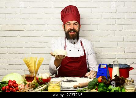 Cuoco con il viso allegro in uniforme di borgogna siede a tavola con verdure e utensili da cucina. L'uomo con la barba tiene l'impasto impastato su sfondo bianco in mattoni. Chef prepara l'impasto. Concetto di cucina professionale Foto Stock