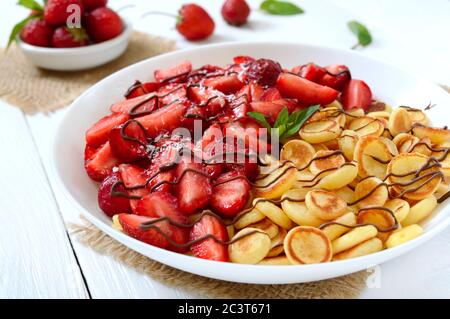 Frittelle di cereali in un recipiente con fragole fresche e noci su sfondo bianco di legno. Colazione gustosa e alla moda. Mini-frittelle olandesi biologiche. Foto Stock
