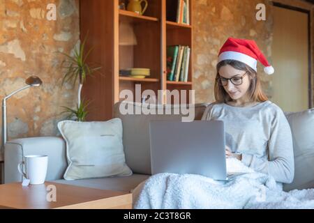 Donna giovane sorridente con cappello Santa guardando un film seduto sul divano a casa di fronte a un computer portatile Foto Stock