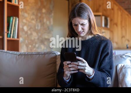 Giovane donna confuso bella bruna scrivere su smartphone seduta sul divano a casa con maglione nero Foto Stock