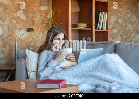 Bella giovane donna sorridente seduto sul divano a casa guardando lo schermo del computer portatile e bevendo un tè. Indossa occhiali e lunghi capelli marroni Foto Stock