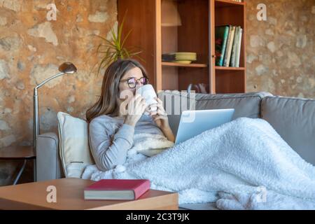 Donna giovane e graziosa, seduta sul divano di casa, guardando lo schermo del computer portatile e bevendo un tè. Indossa occhiali e lunghi capelli marroni. Foto Stock