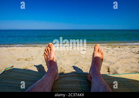 Le gambe dell'uomo sono al sole, distendersi spensierati su un tappetino vicino alla costa, sulla spiaggia pubblica. Foto Stock